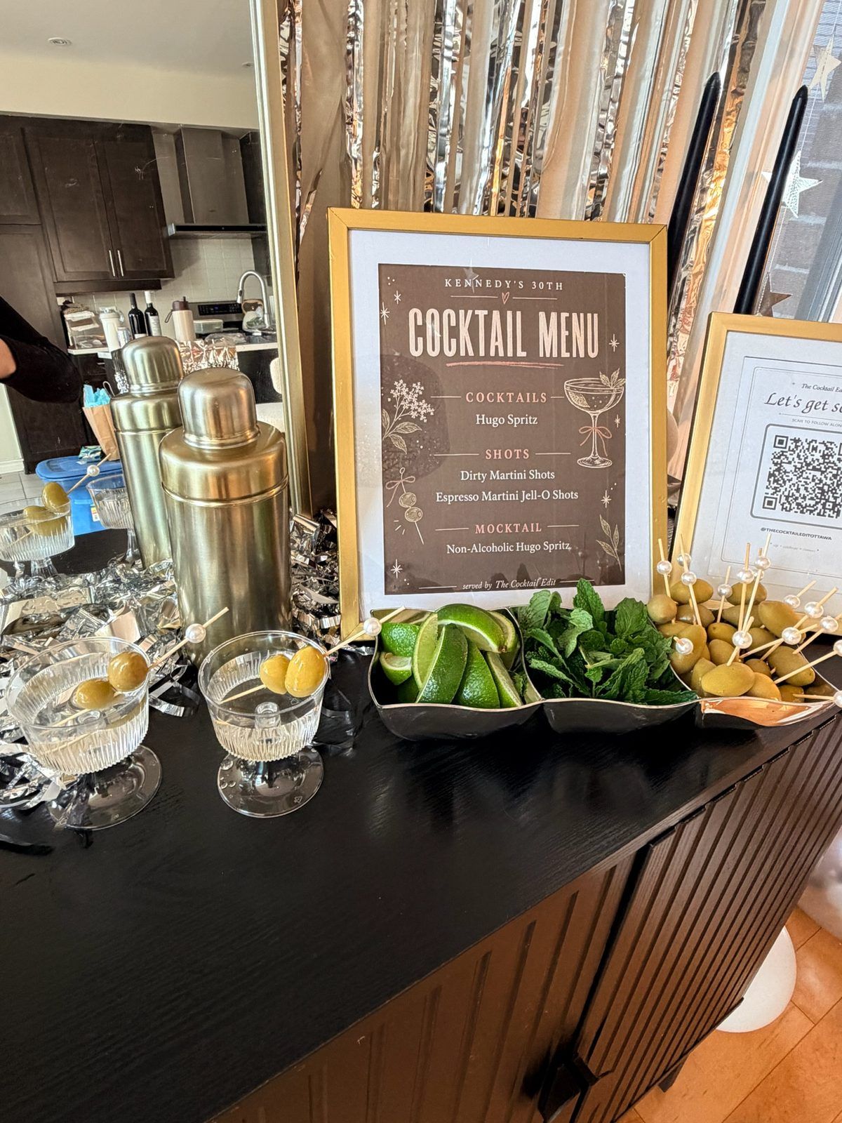 A framed cocktail menu beside a shaker and fresh garnishes