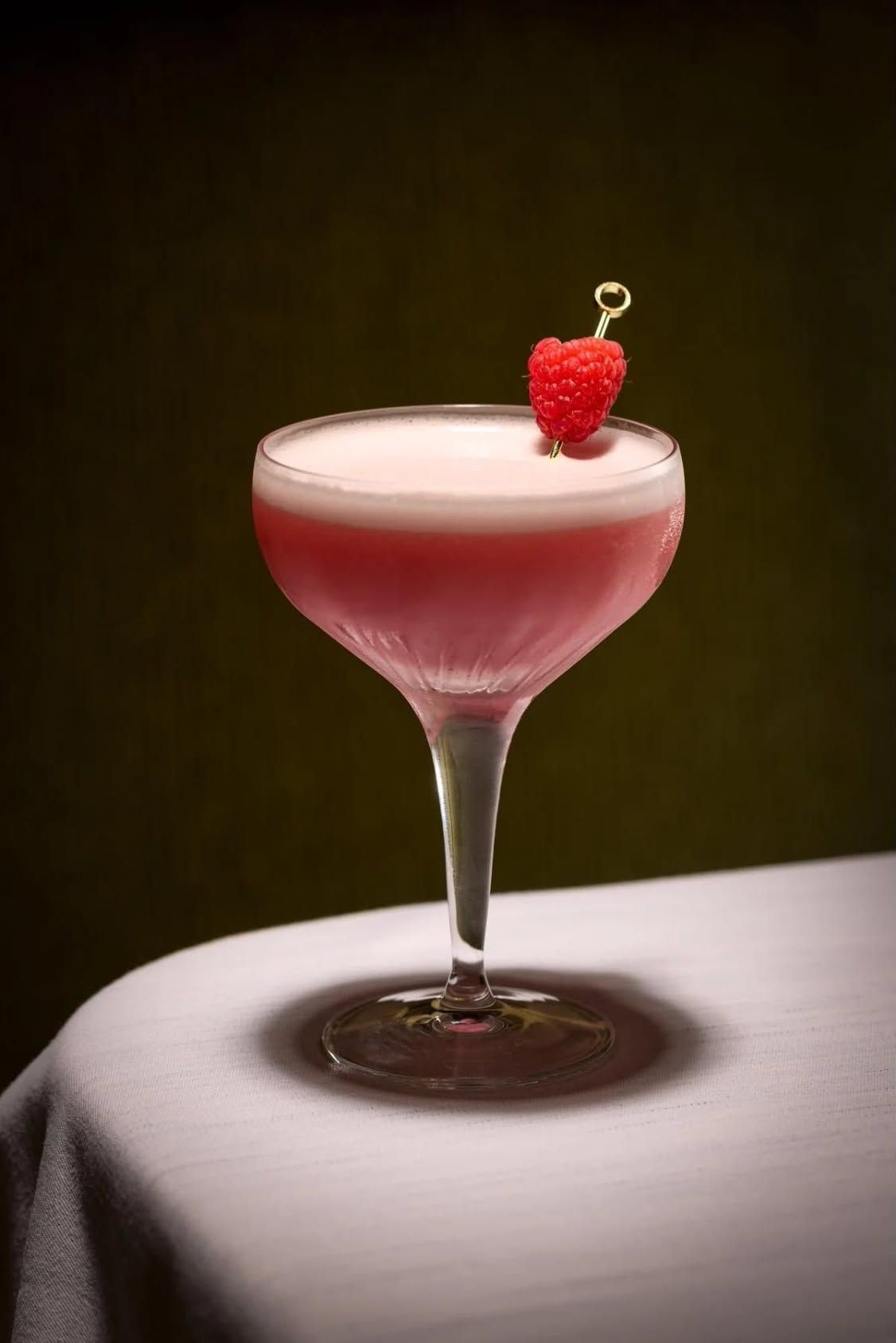 A pink cocktail with a foam top and a raspberry garnish in a coupe glass